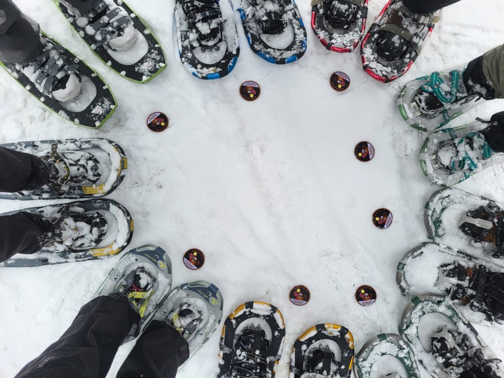 Snowshoe badge hikers in a tight circle with their new Snowshoe Badges on the snow at their feet