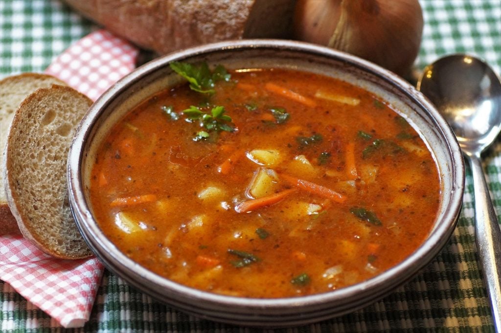 Bowl of steaming goulash soup with crusty bread, onion, spoon and napkin on checked tablecloth